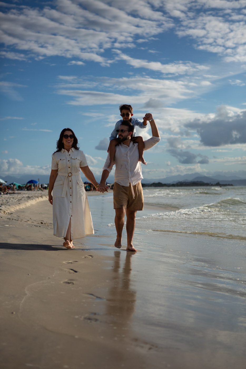 Família caminhando de mãos dadas na beira da praia, com o filho nos ombros do pai