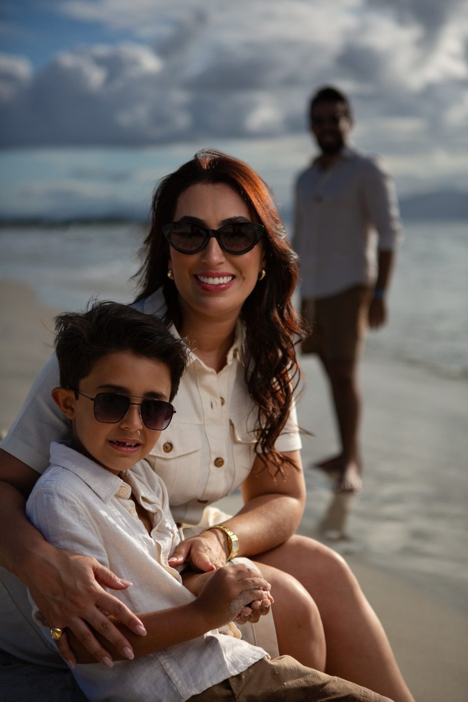 Mãe abraçando o filho na beira da praia, com o pai ao fundo, em tons claros