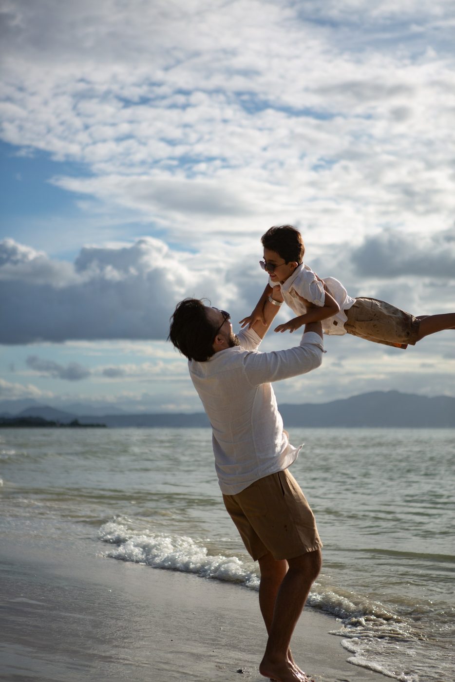 Pai levantando o filho em frente ao mar, com nuvens e luz suave ao fundo